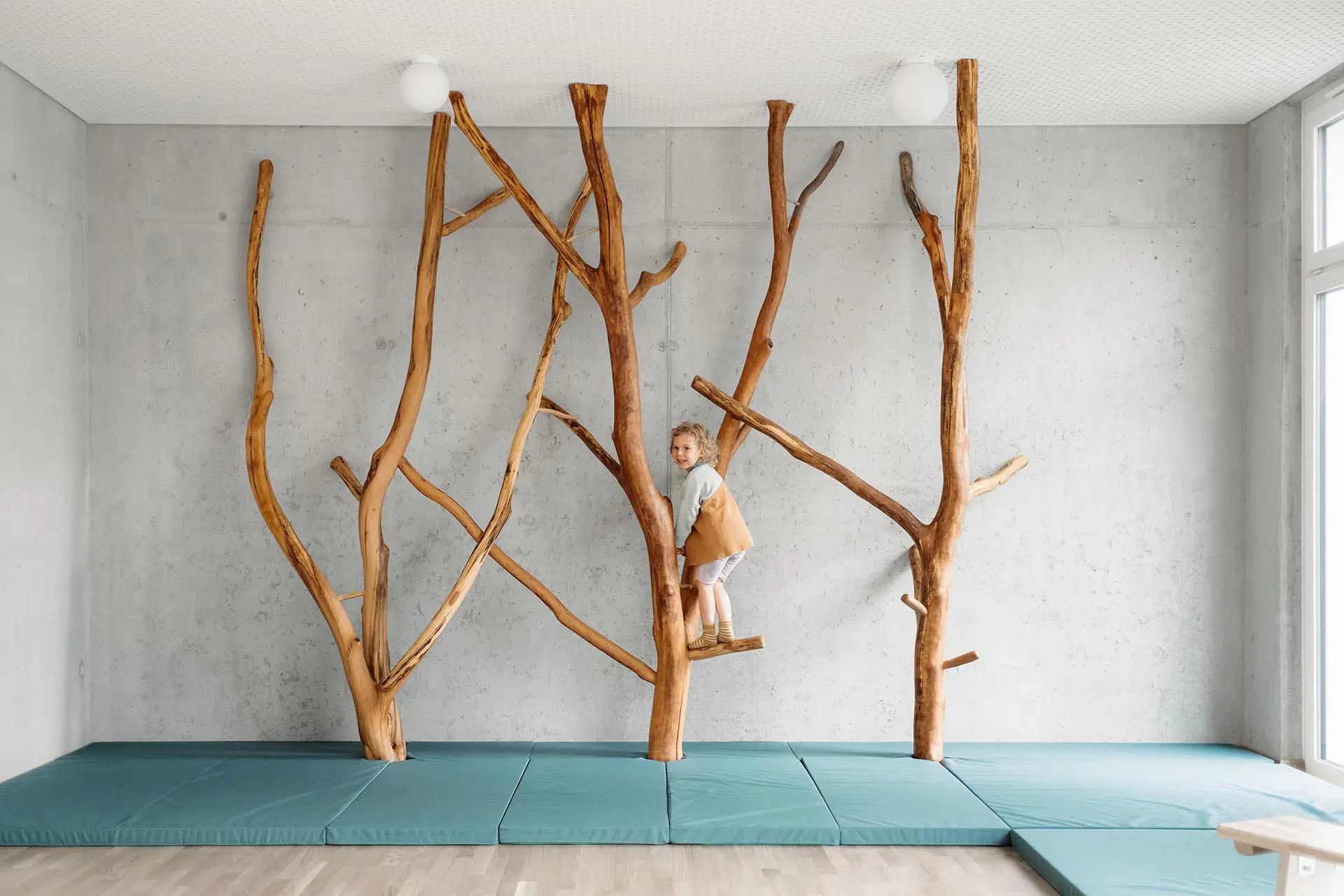 Kindergartenmöbel und Raumgestaltung von papoq: Bewegungsraum einer Kita mit einem Kind, das auf Kletterbäumen klettert, während Sicherheit durch einen Fallschutz gewährleistet ist.