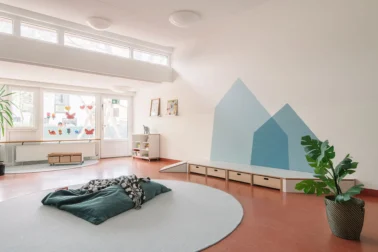 Gruppenraum mit Spielpodest Kindergartenmöbel und Raumgestaltung: Bauchecke mit Spielpodest und herausziehbaren Rollkisten für Spielsachen, Wanddeko in Form einer Baumkrone für naturnahe Atmosphäre in der Kita.