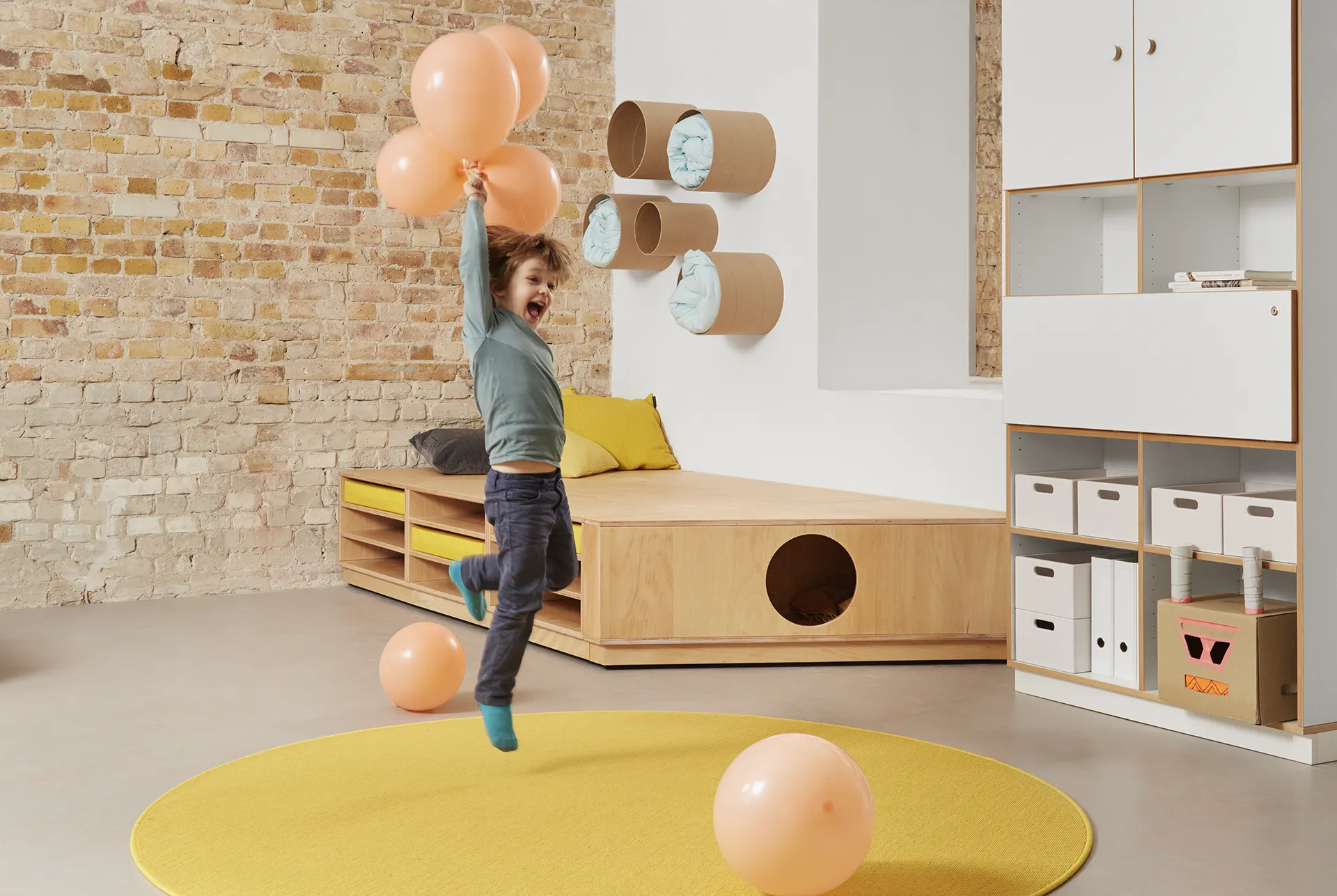 Glückliches Kind mit Luftballons in der Hand vor einem Matratzenpodest, Röhrenregalen und einem Schrank von Papoq – nachhaltige und kindgerechte Möbel für Kindergärten und Horte.
